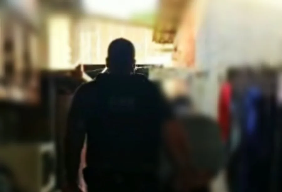 Suspeito de roubo a idoso em Criciúma é preso no Sul Imagem desfocada mostra as costas de um policial civil, vestindo camiseta preta com inscrições brancas, em um ambiente interno durante uma operação.