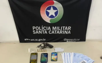 Suspeito é detido com arma e cocaína em Balneário Gaivota Sobre uma mesa branca, estão dispostos itens apreendidos em uma operação policial: um revólver preto, cinco munições, uma pequena porção de cocaína em plástico transparente, três smartphones, diversas cédulas de Real e um maço de folhas de cheque. Ao fundo, uma bandeira preta da Polícia Militar de Santa Catarina com o brasão oficial centralizado.