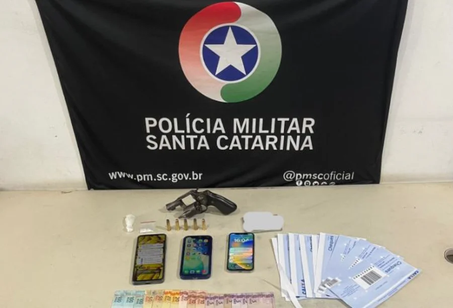 Sobre uma mesa branca, estão dispostos itens apreendidos em uma operação policial: um revólver preto, cinco munições, uma pequena porção de cocaína em plástico transparente, três smartphones, diversas cédulas de Real e um maço de folhas de cheque. Ao fundo, uma bandeira preta da Polícia Militar de Santa Catarina com o brasão oficial centralizado.