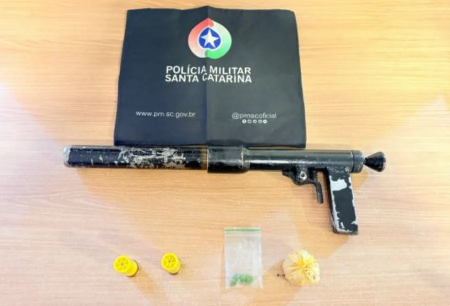 Espingarda artesanal calibre 12, munições, comprimidos de ecstasy e pedra de crack apreendidos pela Polícia Militar de Araranguá, dispostos sobre uma mesa com uma bolsa da PMSC ao fundo.