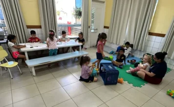 Timbé do Sul inicia Colônia de Férias para 75 crianças Crianças pequenas realizam atividades em uma sala de aula ampla; um grupo está sentado em uma mesa branca desenhando, enquanto outras brincam sobre um tapete de EVA colorido no chão, acompanhadas por uma monitora.