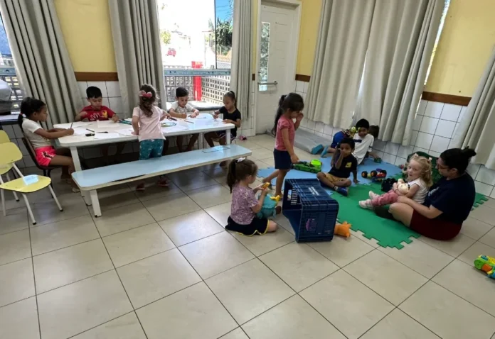 Timbé do Sul inicia Colônia de Férias para 75 crianças Crianças pequenas realizam atividades em uma sala de aula ampla; um grupo está sentado em uma mesa branca desenhando, enquanto outras brincam sobre um tapete de EVA colorido no chão, acompanhadas por uma monitora.