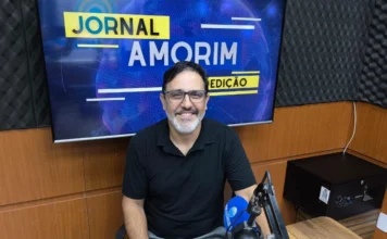 Tina Flores: Trajetória de fé e negócios em Sombrio otografia em plano médio de Ricardo Santana Eugenio, um homem de pele clara, barba grisalha aparada e óculos de grau, sorrindo cordialmente. Ele está sentado à bancada de madeira do estúdio da Rádio 102.9 Amorim FM. À sua frente, destaca-se um microfone profissional articulado. Ao fundo, um monitor exibe a identidade visual do "Jornal Amorim 1ª Edição" em tons de azul e amarelo. A imagem transmite um ambiente profissional e acolhedor de entrevista jornalística.