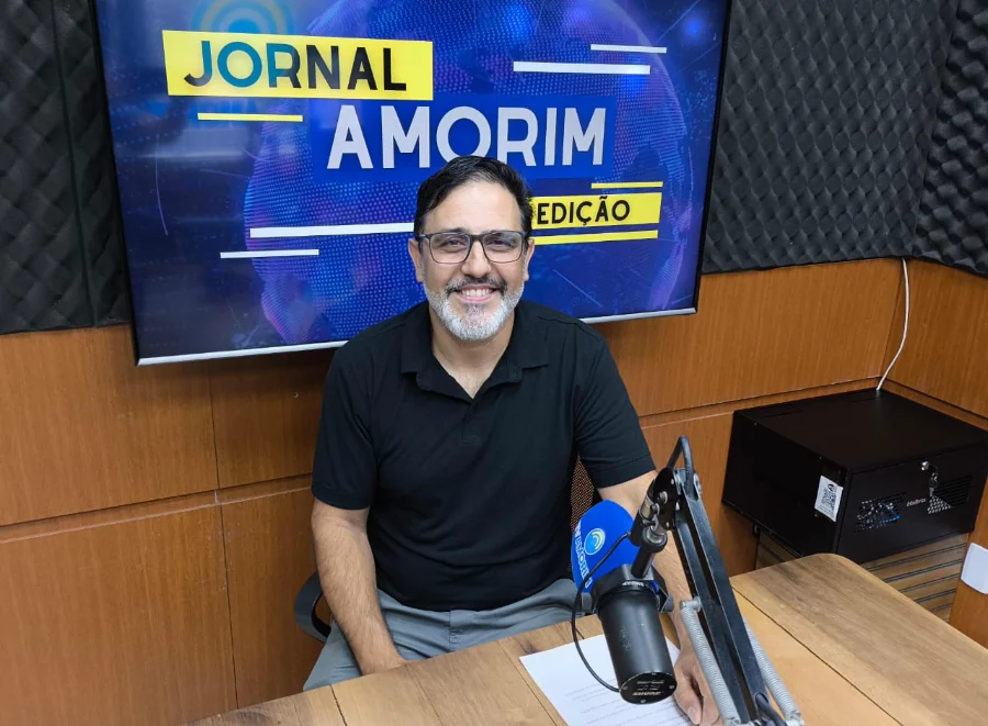 Tina Flores Trajetória de fé e negócios em Sombrio otografia em plano médio de Ricardo Santana Eugenio, um homem de pele clara, barba grisalha aparada e óculos de grau, sorrindo cordialmente. Ele está sentado à bancada de madeira do estúdio da Rádio 102.9 Amorim FM. À sua frente, destaca-se um microfone profissional articulado. Ao fundo, um monitor exibe a identidade visual do