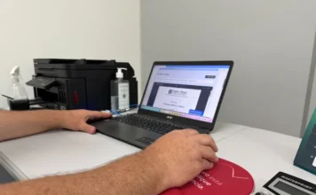 Turvo moderniza sistema digital e prorroga prazos de taxas Fotografia focada em um par de braços e mãos masculinas operando um notebook preto sobre uma mesa branca. Na tela do computador, vê-se um documento do Diário Oficial. Ao lado, encontram-se uma impressora multifuncional preta, álcool em gel e uma calculadora Masterprint branca. A imagem ilustra a modernização dos sistemas digitais em Turvo.