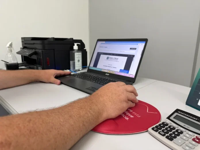 Turvo moderniza sistema digital e prorroga prazos de taxas Fotografia focada em um par de braços e mãos masculinas operando um notebook preto sobre uma mesa branca. Na tela do computador, vê-se um documento do Diário Oficial. Ao lado, encontram-se uma impressora multifuncional preta, álcool em gel e uma calculadora Masterprint branca. A imagem ilustra a modernização dos sistemas digitais em Turvo.