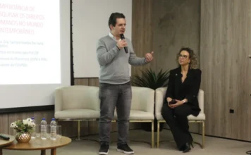 Unesc abre inscrições para o primeiro Doutorado em Direito O coordenador do PPGD da Unesc, Reginaldo de Souza Vieira, fala ao microfone durante um evento de apresentação do novo Doutorado em Direito. Ele está em pé, vestindo suéter cinza, enquanto uma colega o observa sentada ao lado. Ao fundo, um telão exibe o tema "Importância de pesquisar os Direitos Humanos no mundo contemporâneo.