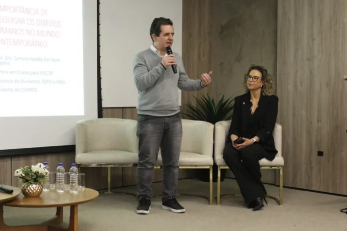 Unesc abre inscrições para o primeiro Doutorado em Direito O coordenador do PPGD da Unesc, Reginaldo de Souza Vieira, fala ao microfone durante um evento de apresentação do novo Doutorado em Direito. Ele está em pé, vestindo suéter cinza, enquanto uma colega o observa sentada ao lado. Ao fundo, um telão exibe o tema "Importância de pesquisar os Direitos Humanos no mundo contemporâneo.
