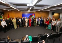 Unesc e China discutem parcerias em Florianópolis Fotografia em plano aberto mostrando um grupo de aproximadamente 30 pessoas, entre homens e mulheres em trajes formais, reunidos em uma sala de conferências do Governo de Santa Catarina. Eles estão posicionados em semicírculo para uma foto oficial. Ao fundo, uma tela exibe a identidade visual do Governo do Estado e, nas laterais, as bandeiras do Brasil e de Santa Catarina. No primeiro plano, é possível ver algumas cadeiras e pastas da Unesc sobre o chão.