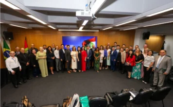 Unesc e China discutem parcerias em Florianópolis Fotografia em plano aberto mostrando um grupo de aproximadamente 30 pessoas, entre homens e mulheres em trajes formais, reunidos em uma sala de conferências do Governo de Santa Catarina. Eles estão posicionados em semicírculo para uma foto oficial. Ao fundo, uma tela exibe a identidade visual do Governo do Estado e, nas laterais, as bandeiras do Brasil e de Santa Catarina. No primeiro plano, é possível ver algumas cadeiras e pastas da Unesc sobre o chão.