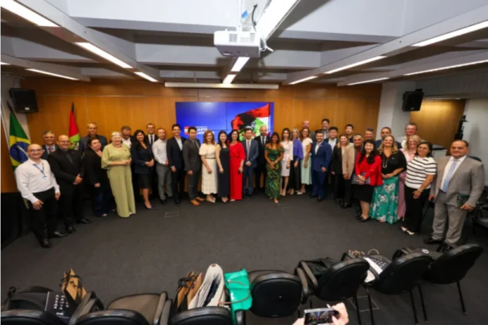 Unesc e China discutem parcerias em Florianópolis Fotografia em plano aberto mostrando um grupo de aproximadamente 30 pessoas, entre homens e mulheres em trajes formais, reunidos em uma sala de conferências do Governo de Santa Catarina. Eles estão posicionados em semicírculo para uma foto oficial. Ao fundo, uma tela exibe a identidade visual do Governo do Estado e, nas laterais, as bandeiras do Brasil e de Santa Catarina. No primeiro plano, é possível ver algumas cadeiras e pastas da Unesc sobre o chão.