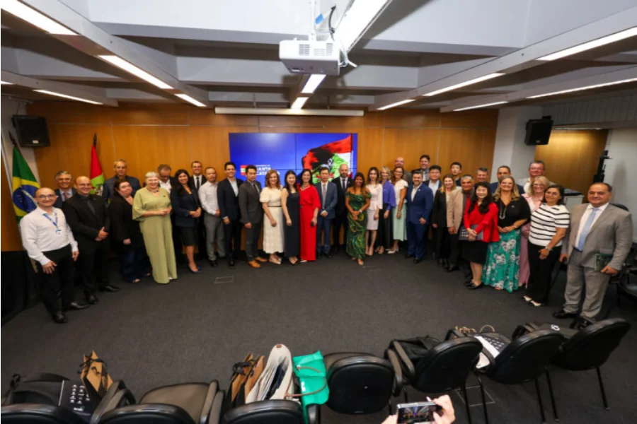 Unesc e China discutem parcerias em Florianópolis Fotografia em plano aberto mostrando um grupo de aproximadamente 30 pessoas, entre homens e mulheres em trajes formais, reunidos em uma sala de conferências do Governo de Santa Catarina. Eles estão posicionados em semicírculo para uma foto oficial. Ao fundo, uma tela exibe a identidade visual do Governo do Estado e, nas laterais, as bandeiras do Brasil e de Santa Catarina. No primeiro plano, é possível ver algumas cadeiras e pastas da Unesc sobre o chão.