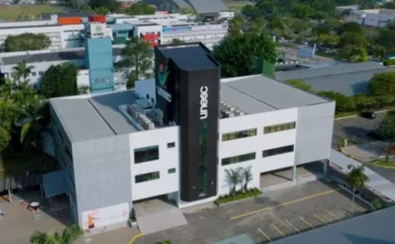 Unesc encerra inscrições para Universidade Gratuita hoje Vista aérea do prédio da Unesc, com arquitetura moderna em tons de branco e cinza e uma torre preta central exibindo o logotipo da universidade em branco. O entorno mostra áreas arborizadas e o estacionamento do campus, local onde funciona a Central de Atendimento ao Acadêmico (Centac) para orientações sobre o Programa Universidade Gratuita.