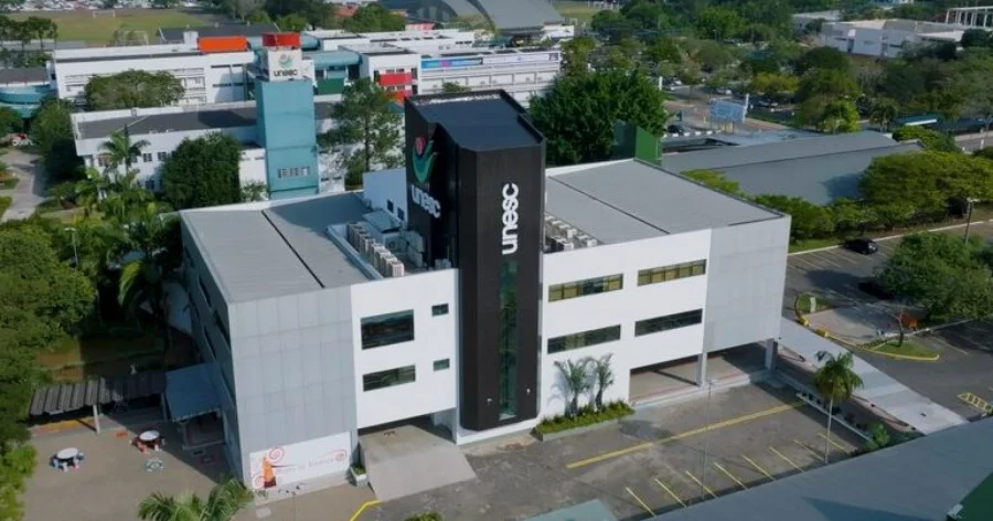Unesc encerra inscrições para Universidade Gratuita hoje Vista aérea do prédio da Unesc, com arquitetura moderna em tons de branco e cinza e uma torre preta central exibindo o logotipo da universidade em branco. O entorno mostra áreas arborizadas e o estacionamento do campus, local onde funciona a Central de Atendimento ao Acadêmico (Centac) para orientações sobre o Programa Universidade Gratuita.