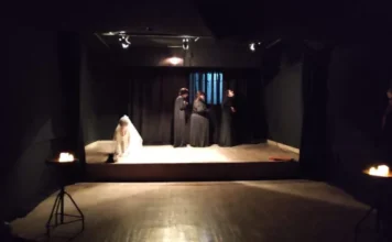 Unesc lança Licenciatura em Artes Cênicas no Sul de SC Fotografia de uma cena teatral em um palco com fundo e cortinas pretas. No centro, três pessoas vestem túnicas longas escuras em uma composição estática. À esquerda, uma pessoa com vestes brancas está agachada no chão sob o foco de luz. A iluminação é pontual, vinda de refletores superiores e de velas posicionadas sobre suportes de ferro nas laterais, criando um ambiente dramático que ilustra as atividades práticas do curso de Artes Cênicas.