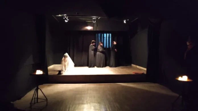 Unesc lança Licenciatura em Artes Cênicas no Sul de SC Fotografia de uma cena teatral em um palco com fundo e cortinas pretas. No centro, três pessoas vestem túnicas longas escuras em uma composição estática. À esquerda, uma pessoa com vestes brancas está agachada no chão sob o foco de luz. A iluminação é pontual, vinda de refletores superiores e de velas posicionadas sobre suportes de ferro nas laterais, criando um ambiente dramático que ilustra as atividades práticas do curso de Artes Cênicas.