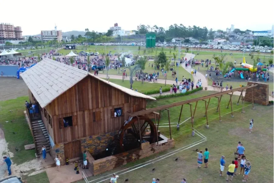 Vista aérea do Parque dos Imigrantes, no Distrito do Rio Maina, local que recebe a ação educativa do Colégio Unesc. A imagem destaca uma grande construção de madeira com uma roda d'água em funcionamento, cercada por caminhos onde diversas pessoas caminham pelo gramado. Ao fundo, é possível observar tendas de eventos e uma área de lazer infantil com um escorregador em formato de avião amarelo.