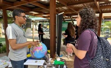 Unesc retoma curso de Geografia para suprir falta de docentes Fotografia de um estande informativo do curso de Geografia da Unesc em um ambiente externo. À esquerda, um homem de óculos e camiseta cinza aponta para um globo terrestre colorido sobre uma mesa. À direita, uma jovem de cabelos cacheados e mochila observa a explicação. A mesa está coberta com uma toalha com o logotipo da Unesc e exibe amostras de rochas, minerais e mapas em relevo 3D, simbolizando a formação prática e a interação com a comunidade.