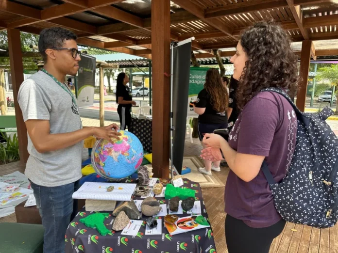 Unesc retoma curso de Geografia para suprir falta de docentes Fotografia de um estande informativo do curso de Geografia da Unesc em um ambiente externo. À esquerda, um homem de óculos e camiseta cinza aponta para um globo terrestre colorido sobre uma mesa. À direita, uma jovem de cabelos cacheados e mochila observa a explicação. A mesa está coberta com uma toalha com o logotipo da Unesc e exibe amostras de rochas, minerais e mapas em relevo 3D, simbolizando a formação prática e a interação com a comunidade.