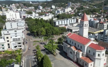Urussanga abre 441 empresas e supera saldo de 2024 Vista aérea do centro de Urussanga, com destaque para a Igreja Matriz de arquitetura clara e telhado vermelho. Ao redor, veem-se ruas arborizadas com palmeiras, prédios baixos e a paisagem urbana da cidade sob a luz do sol.