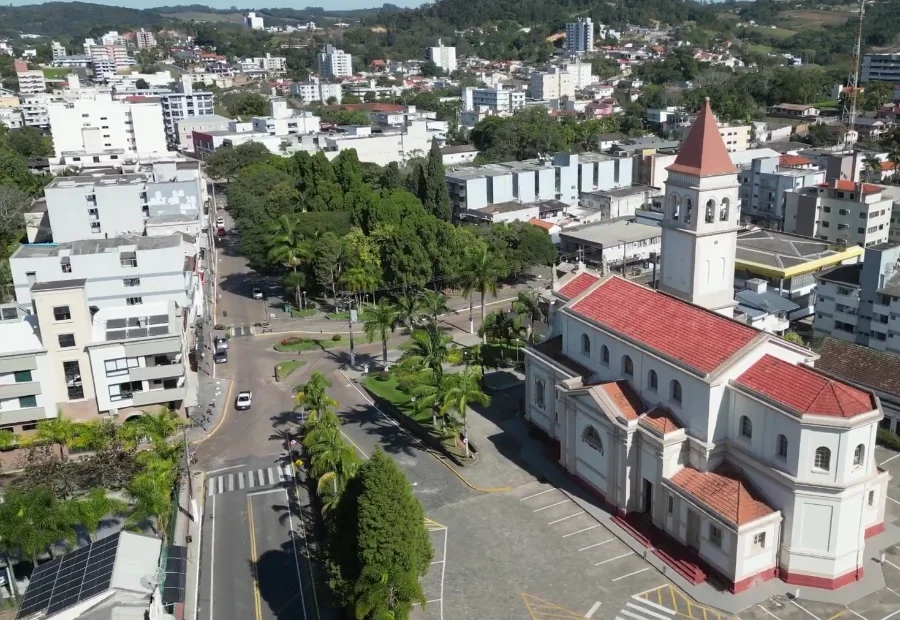Vista aérea do centro de Urussanga, com destaque para a Igreja Matriz de arquitetura clara e telhado vermelho. Ao redor, veem-se ruas arborizadas com palmeiras, prédios baixos e a paisagem urbana da cidade sob a luz do sol.