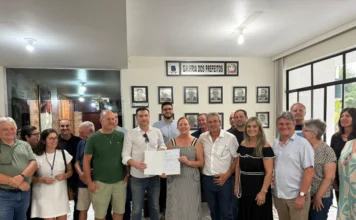Urussanga autoriza asfalto na Rodovia da Imigração Italiana A imagem mostra um grupo de cerca de vinte pessoas reunidas em um saguão da prefeitura de Urussanga, com uma parede ao fundo contendo retratos e a inscrição "GALERIA DOS PREFEITOS". No centro, a prefeita Stela de Agostin Talamini (mulher loira com vestido listrado) e um homem de camisa branca seguram juntos um documento aberto, que é a ordem de serviço assinada. Eles estão cercados por outras autoridades e membros da comunidade, todos posando para a foto.