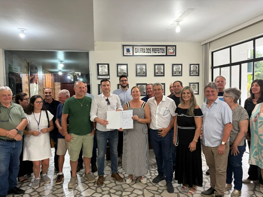 A imagem mostra um grupo de cerca de vinte pessoas reunidas em um saguão da prefeitura de Urussanga, com uma parede ao fundo contendo retratos e a inscrição 