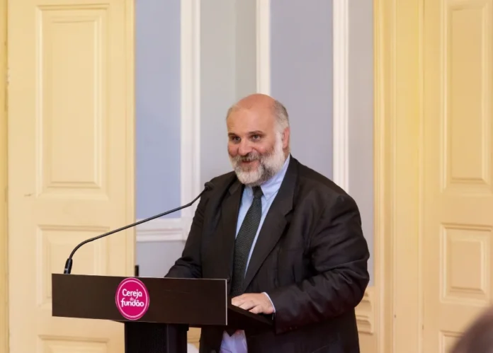 V Fórum da Abadeus traz palestrante de Portugal Paulo Alexandre Bernardo Fernandes, presidente da Câmara Municipal do Fundão em Portugal e palestrante confirmado do V Fórum da Abadeus, aparece discursando em um púlpito. Ele é um homem de pele clara, com barba e cabelos grisalhos, vestindo terno cinza escuro, camisa azul clara e gravata. No púlpito, há um selo circular com a inscrição "Cereja do Fundão".