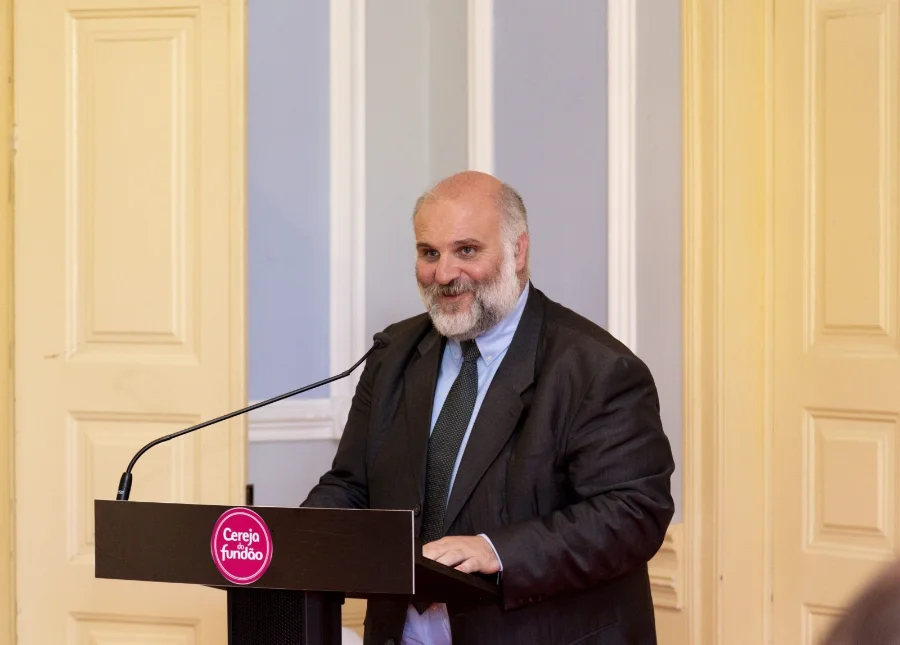 V Fórum da Abadeus traz palestrante de Portugal Paulo Alexandre Bernardo Fernandes, presidente da Câmara Municipal do Fundão em Portugal e palestrante confirmado do V Fórum da Abadeus, aparece discursando em um púlpito. Ele é um homem de pele clara, com barba e cabelos grisalhos, vestindo terno cinza escuro, camisa azul clara e gravata. No púlpito, há um selo circular com a inscrição