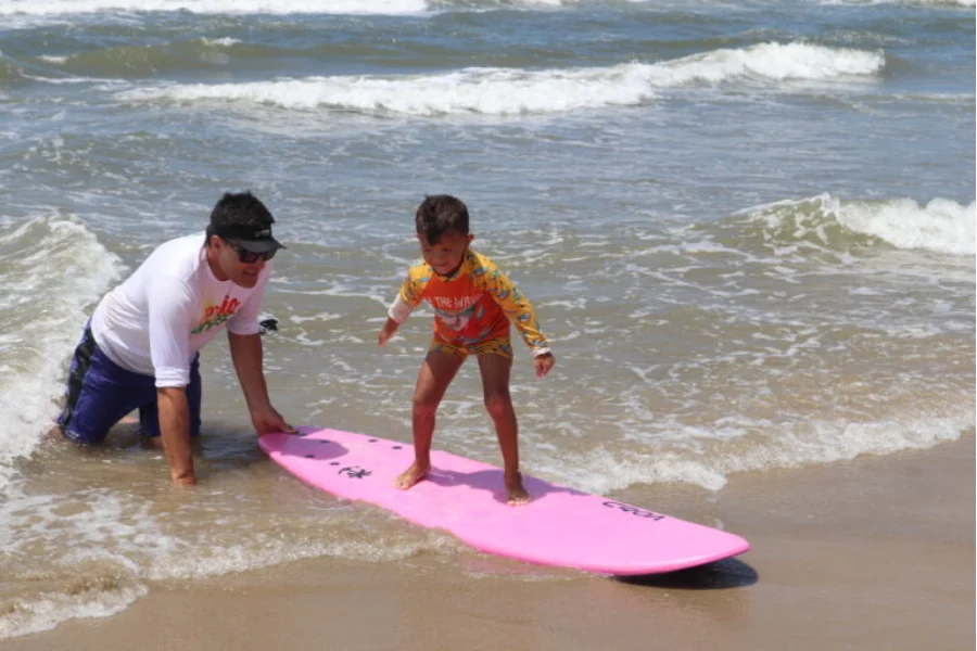 Instrutor auxilia um menino a ficar em pé em uma prancha de surfe rosa no mar durante uma aula do projeto Verão Unesc, no Balneário Rincão.