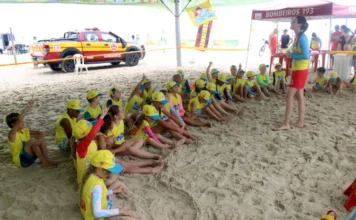 Verão Unesc movimenta orla do Rincão com serviços e lazer Crianças participantes do Programa Golfinho sentadas na areia da praia ouvindo orientações de uma instrutora, com viatura e tenda do Corpo de Bombeiros ao fundo, durante ação do Verão Unesc no Balneário Rincão.