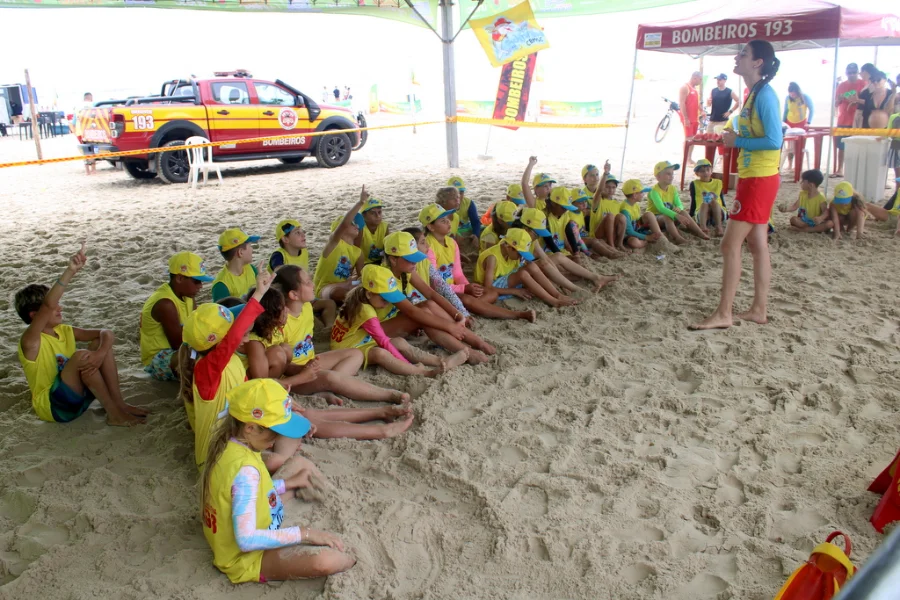 Verão Unesc movimenta orla do Rincão com serviços e lazer Crianças participantes do Programa Golfinho sentadas na areia da praia ouvindo orientações de uma instrutora, com viatura e tenda do Corpo de Bombeiros ao fundo, durante ação do Verão Unesc no Balneário Rincão.