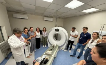 Vigilância Sanitária de SC divulga balanço positivo de 2025 Grupo de cerca de dez profissionais, incluindo um homem de jaleco branco que fala gesticulando, reunidos em uma sala de exames médicos com um grande equipamento de tomografia computadorizada ao fundo. Sobre uma maca, há diversos instrumentos técnicos. A cena ilustra as ações de fiscalização e capacitação em alta complexidade descritas no balanço da DIVS.