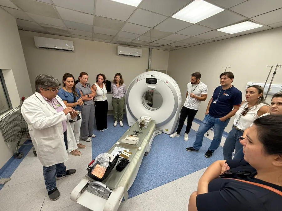 Grupo de cerca de dez profissionais, incluindo um homem de jaleco branco que fala gesticulando, reunidos em uma sala de exames médicos com um grande equipamento de tomografia computadorizada ao fundo. Sobre uma maca, há diversos instrumentos técnicos. A cena ilustra as ações de fiscalização e capacitação em alta complexidade descritas no balanço da DIVS.