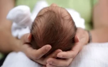 Zika: risco de internação por epilepsia é 34 vezes maior Mãos de adulto segurando com cuidado a cabeça de um recém-nascido de cabelos escuros, ilustrando a vulnerabilidade e a necessidade de atenção neurológica em bebês, tema central do estudo da Fiocruz sobre a Síndrome Congênita do Zika e o risco de epilepsia.