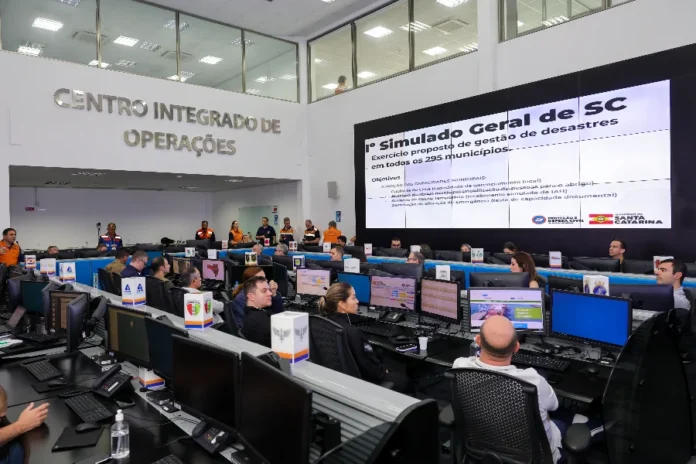 1º Exercício Geral de Gestão de Desastres – MAIO 2025 FOTO Roberto Zacarias SecomGOVSC 2 Uma foto ampla do interior do Centro Integrado de Operações em Santa Catarina, repleto de dezenas de profissionais de defesa civil sentados em fileiras de estações de trabalho monitorando computadores. No fundo da sala, uma grande tela de vídeo exibe o título "1º Simulado Geral de SC" e o subtítulo "Exercício proposto de gestão de desastres em todos os 295 municípios". A tela também detalha objetivos específicos como "Gabinete de Crise (capacidade de gerenciamento local)" e "Abertura de abrigo municipal", além de mostrar os logotipos da Defesa Civil de Santa Catarina e do Governo do Estado. Letras grandes de metal na parede à esquerda dizem "CENTRO INTEGRADO DE OPERAÇÕES". Esta imagem ilustra o primeiro exercício geral de gestão de desastres do estado, que estabeleceu o recorde de participação nacional que o segundo simulado anunciado na matéria superará.