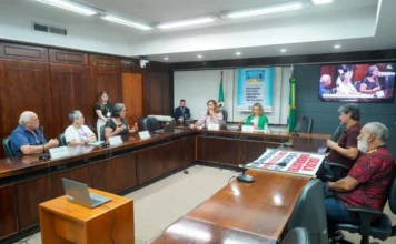 ALRS aprova novos projetos de cultura e inovação no RS Fotografia colorida da reunião da Comissão de Educação, Cultura, Desporto, Ciência e Tecnologia na Assembleia Legislativa do Rio Grande do Sul. Em uma sala com painéis de madeira, parlamentares e representantes da comunidade estão sentados em torno de uma grande mesa em formato de "U". No centro, ao fundo, um banner azul identifica a comissão ao lado das bandeiras oficiais. À esquerda, uma representante da comunidade fala ao microfone, enquanto sobre a mesa, em destaque, há um cartaz com os dizeres "Escola Embratel". Um monitor de TV à direita exibe a transmissão simultânea do encontro.