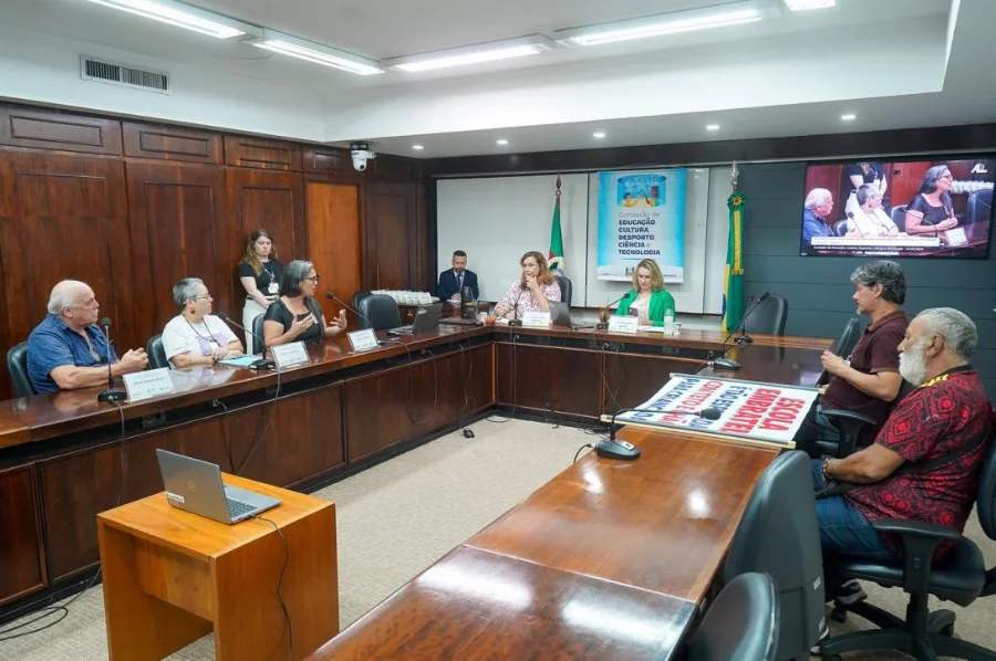 Fotografia colorida da reunião da Comissão de Educação, Cultura, Desporto, Ciência e Tecnologia na Assembleia Legislativa do Rio Grande do Sul. Em uma sala com painéis de madeira, parlamentares e representantes da comunidade estão sentados em torno de uma grande mesa em formato de 