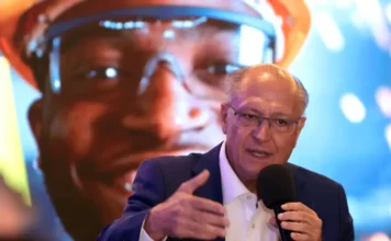 Acordo Mercosul-UE deve entrar em vigor em maio Foto em ângulo médio de Geraldo Alckmin, vice-presidente e ministro do Desenvolvimento, Indústria, Comércio e Serviços, de terno azul e óculos, falando em um microfone com a mão direita estendida para enfatizar suas palavras. Ele está em um evento, falando sobre o acordo comercial Mercosul-UE. Ao fundo, uma grande tela projeta um close-up do rosto de um trabalhador negro usando capacete de segurança laranja e óculos de proteção, simbolizando os setores industriais e de trabalho que podem ser impactados pelo acordo. A iluminação é de palco com efeitos de luz desfocados.