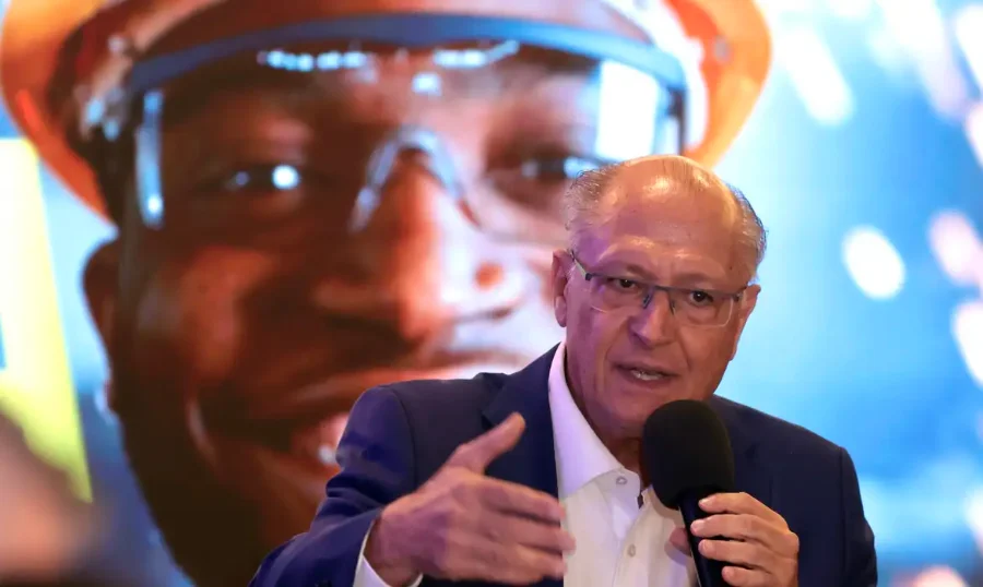 Foto em ângulo médio de Geraldo Alckmin, vice-presidente e ministro do Desenvolvimento, Indústria, Comércio e Serviços, de terno azul e óculos, falando em um microfone com a mão direita estendida para enfatizar suas palavras. Ele está em um evento, falando sobre o acordo comercial Mercosul-UE. Ao fundo, uma grande tela projeta um close-up do rosto de um trabalhador negro usando capacete de segurança laranja e óculos de proteção, simbolizando os setores industriais e de trabalho que podem ser impactados pelo acordo. A iluminação é de palco com efeitos de luz desfocados.