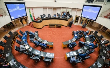 Alesc aprova desconto no IPVA para bons motoristas Vista ampla e elevada do Plenário da Assembleia Legislativa de Santa Catarina (Alesc) durante a sessão ordinária em que foram aprovados projetos como o desconto no IPVA para bons motoristas e a equiparação de direitos para pacientes com hipertensão pulmonar. Os deputados estão sentados em suas bancadas dispostas em semicírculo sobre um tapete vermelho, com telões ao fundo exibindo a pauta de votação.