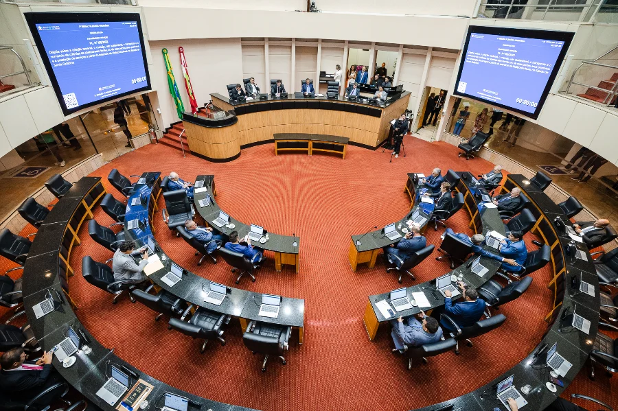 Vista ampla e elevada do Plenário da Assembleia Legislativa de Santa Catarina (Alesc) durante a sessão ordinária em que foram aprovados projetos como o desconto no IPVA para bons motoristas e a equiparação de direitos para pacientes com hipertensão pulmonar. Os deputados estão sentados em suas bancadas dispostas em semicírculo sobre um tapete vermelho, com telões ao fundo exibindo a pauta de votação.