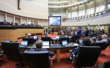 Alesc aprova fim dos fogos de estampido em Santa Catarina Vista ampla do Plenário da Assembleia Legislativa de Santa Catarina (Alesc) durante sessão ordinária. Parlamentares estão sentados em suas bancadas organizadas em semicírculo, com notebooks abertos. Ao centro, um grande monitor exibe a transmissão ao vivo da fala de um dos deputados. O ambiente possui iluminação clara e arquitetura moderna com detalhes em madeira.