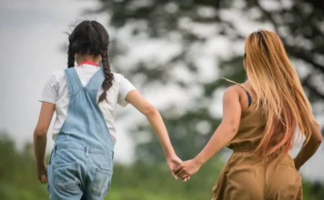 Alesc aprova projeto de apadrinhamento afetivo em SC Mulher e criança caminham de mãos dadas em um gramado ao ar livre, vistas de costas. A imagem simboliza o vínculo, a confiança e o suporte emocional proporcionados pelo programa de apadrinhamento afetivo a crianças e adolescentes em situação de acolhimento.