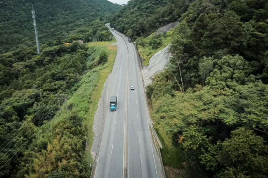 Vista aérea de um trecho sinuoso da rodovia BR-101 cortando a densa vegetação do Morro dos Cavalos, na Grande Florianópolis. A imagem mostra a pista dupla, alguns veículos e encostas íngremes, ilustrando a área considerada de risco que é pauta de debates na Alesc sobre a necessidade de obras de segurança.