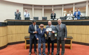 Alesc homenageia os 70 anos da Epagri em sessão especial Sessão especial na Assembleia Legislativa de Santa Catarina (Alesc). Quatro homens estão em pé no centro do auditório com carpete vermelho. O homem idoso, Glauco Olinger, com boné e bengala, recebe uma placa com a ajuda de outros homens de terno. Pessoas na tribuna elevada ao fundo aplaudem a cena.