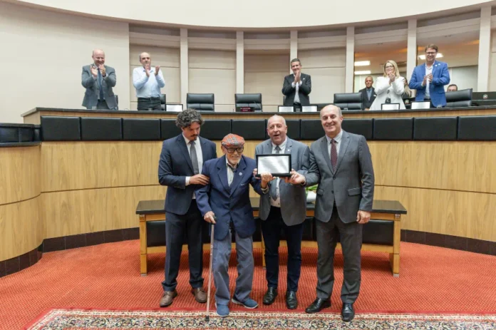 Alesc homenageia os 70 anos da Epagri em sessão especial Sessão especial na Assembleia Legislativa de Santa Catarina (Alesc). Quatro homens estão em pé no centro do auditório com carpete vermelho. O homem idoso, Glauco Olinger, com boné e bengala, recebe uma placa com a ajuda de outros homens de terno. Pessoas na tribuna elevada ao fundo aplaudem a cena.