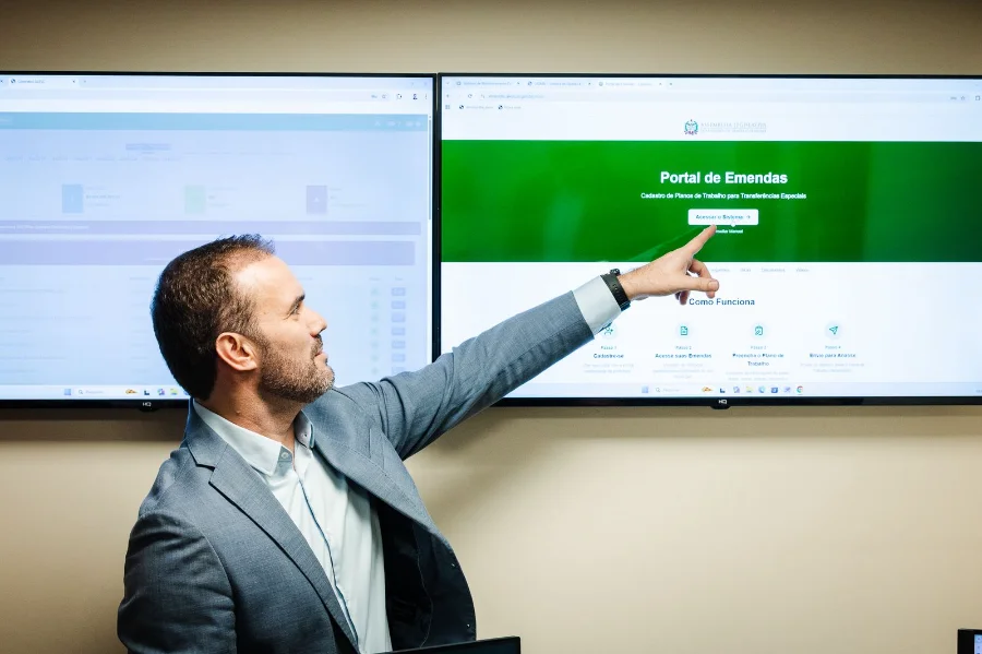 Homem em traje formal aponta para um grande monitor que exibe a interface inicial do Portal de Emendas da Assembleia Legislativa de Santa Catarina. A tela, em tons de verde e branco, destaca as etapas de cadastro e envio de planos de trabalho, simbolizando a digitalização e a transparência no processo de repasse de recursos aos municípios.