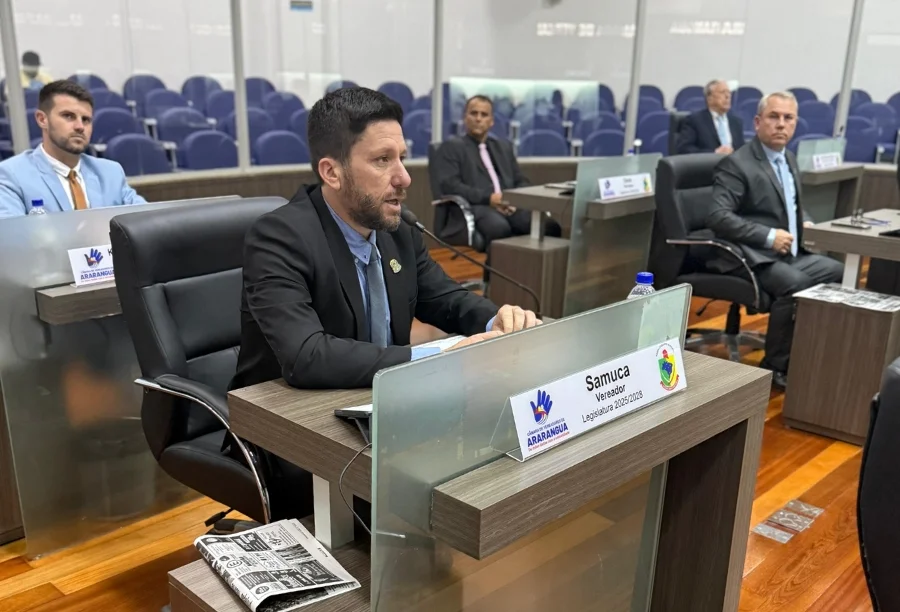 O vereador Samuca discursa na tribuna da Câmara Municipal de Araranguá durante a sessão onde sugeriu a criação da comissão de fiscalização da Quarta Ponte. Ao fundo, outros vereadores estão sentados no plenário.