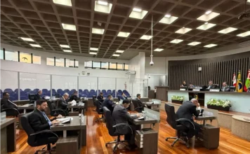 Araranguá aprova projetos de saúde e infraestrutura Fotografia do plenário da Câmara de Vereadores de Araranguá durante a segunda sessão ordinária de 2026. A imagem mostra os parlamentares sentados em suas bancadas individuais de madeira, organizadas em semicírculo, focados em documentos e equipamentos eletrônicos. Ao fundo, destaca-se a mesa diretora sob uma parede com painéis verticais de madeira e as bandeiras oficiais. O ambiente possui iluminação moderna embutida em um teto quadriculado e piso de madeira polida, refletindo o caráter solene da votação de projetos de saúde e infraestrutura.